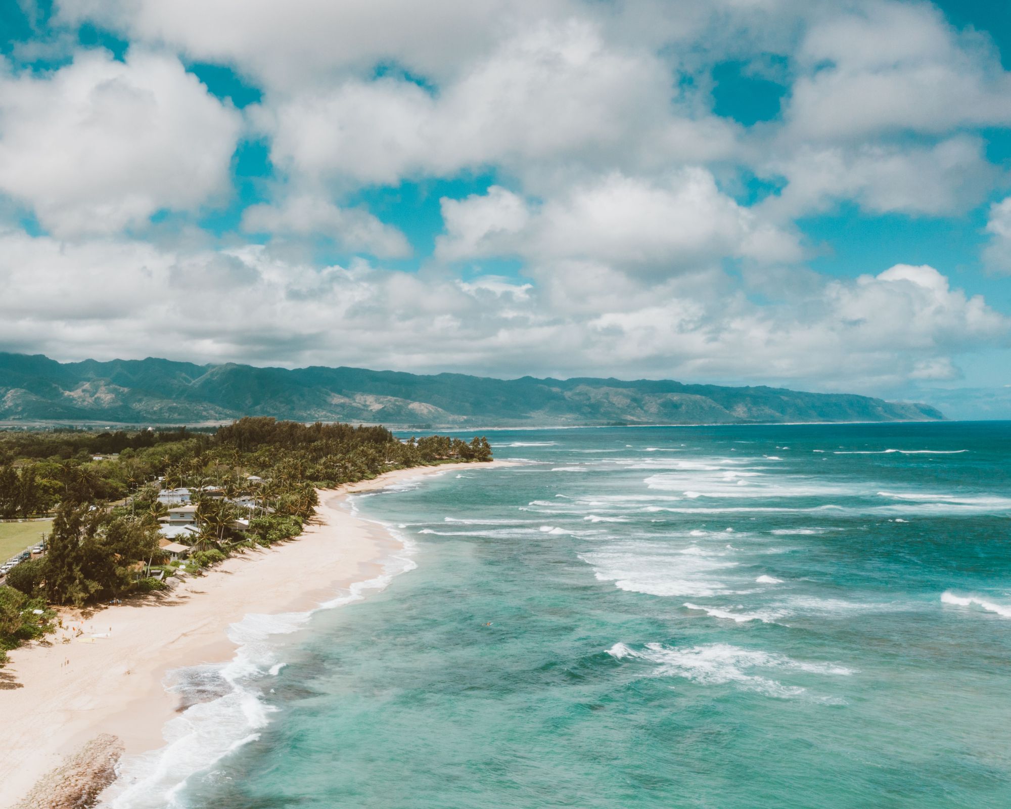 Aerial view of the North Shore of Oahu, with a long beach, green hills and the open ocean
