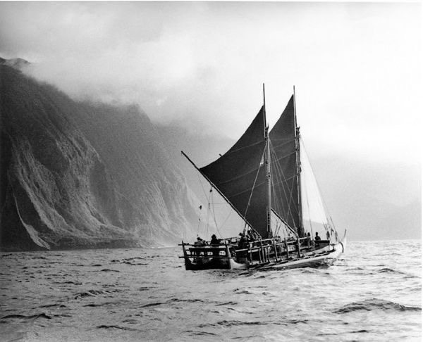 A double-hulled voyaging canoe under sail beneath sea cliffs