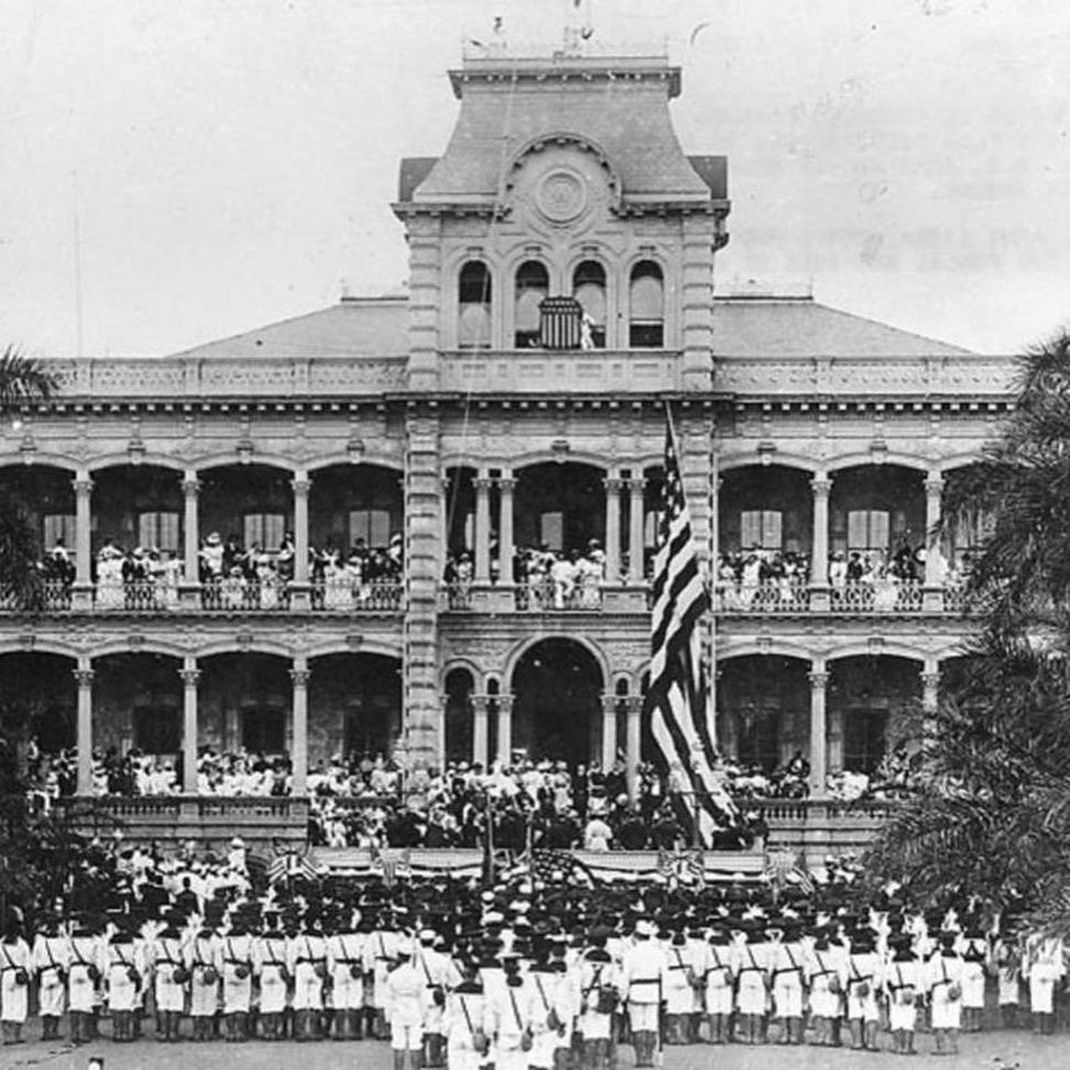 Black-and-white photograph of the annexation ceremony at ʻIolani Palace, with ranks of uniformed troops on the lawn, a large American flag on a pole and crowds on the palace verandas