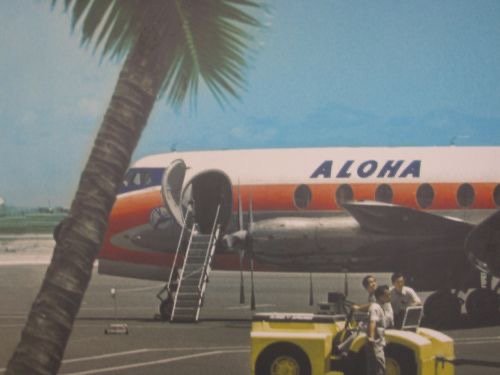 Colour photograph of an Aloha Airlines Vickers Viscount turboprop on the airport ramp with its boarding stairs down, a palm tree in front and a yellow tug beside it, 1960s