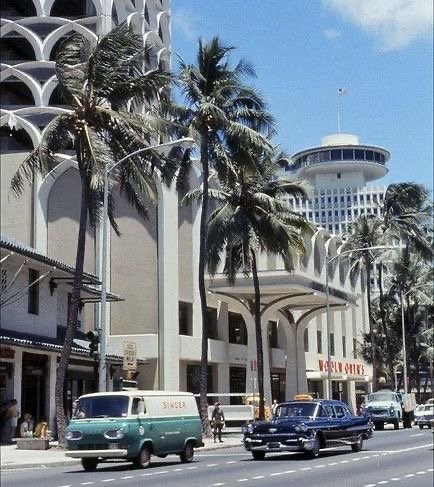 A Waikīkī street in the 1960s with tall palms, a green Singer van and a dark sedan in front of modern hotel towers