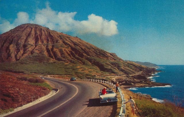 Colour postcard of a white convertible parked on a winding coast road below the reddish slopes of Koko Head, Oʻahu, with the blue ocean on the right, 1950s