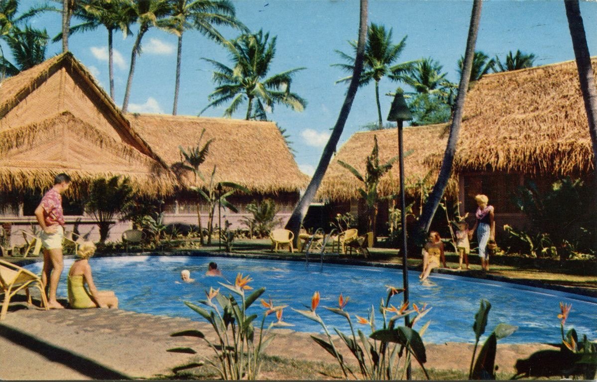 Colour postcard from the 1950s showing a hotel pool surrounded by thatched-roof cottages and palms, with guests in swimwear, a man in an aloha shirt and birds of paradise in the foreground