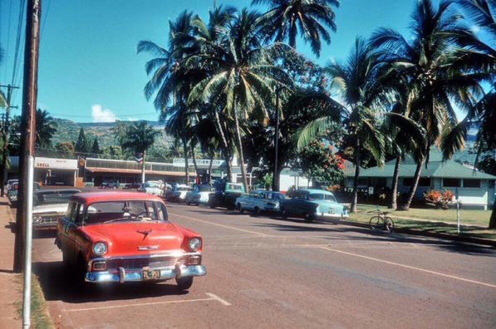 Colour photo of a small-town Hawaiʻi street in the late 1950s, with a red 1956 Chevrolet parked in front, more period cars beyond and tall coconut palms