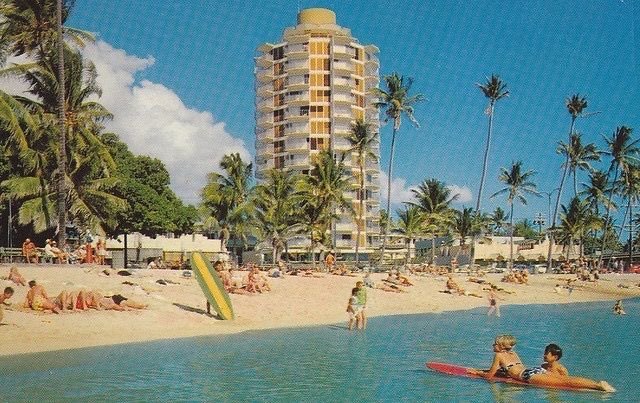 Colour postcard from the 1960s showing a round-fronted high-rise hotel behind a Waikīkī beach lagoon, with palms, sunbathers, a yellow surfboard and two people on a red board in the water