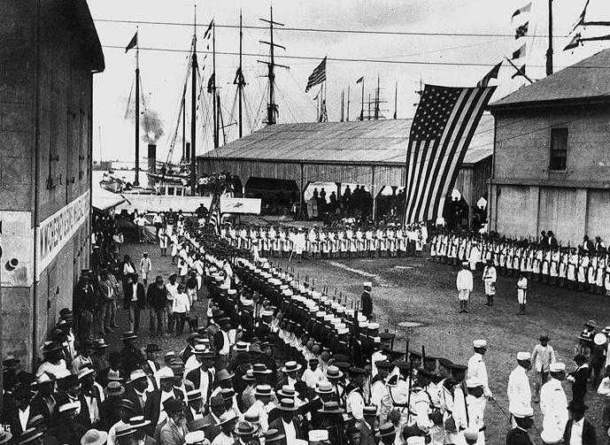 Rows of American sailors in white uniforms on a Honolulu wharf beneath a large US flag, with spectators and ships' masts behind
