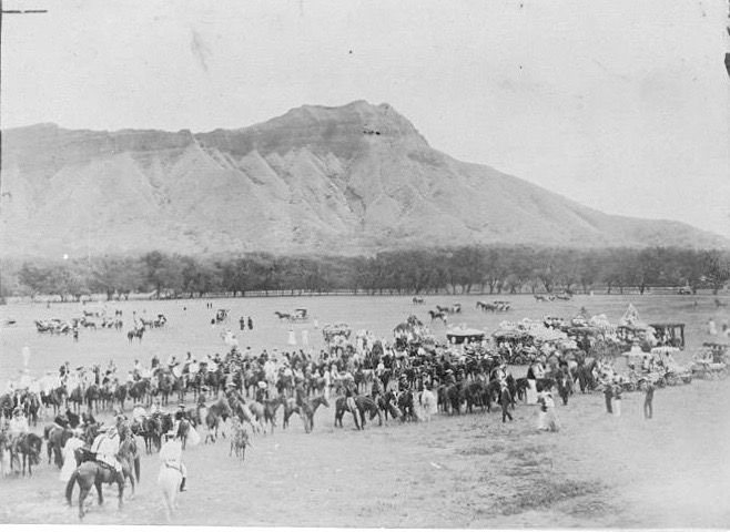 Black-and-white photograph of a large crowd of riders on horseback and carriages gathered on the open lawns of Kapiʻolani Park below Diamond Head, around 1900
