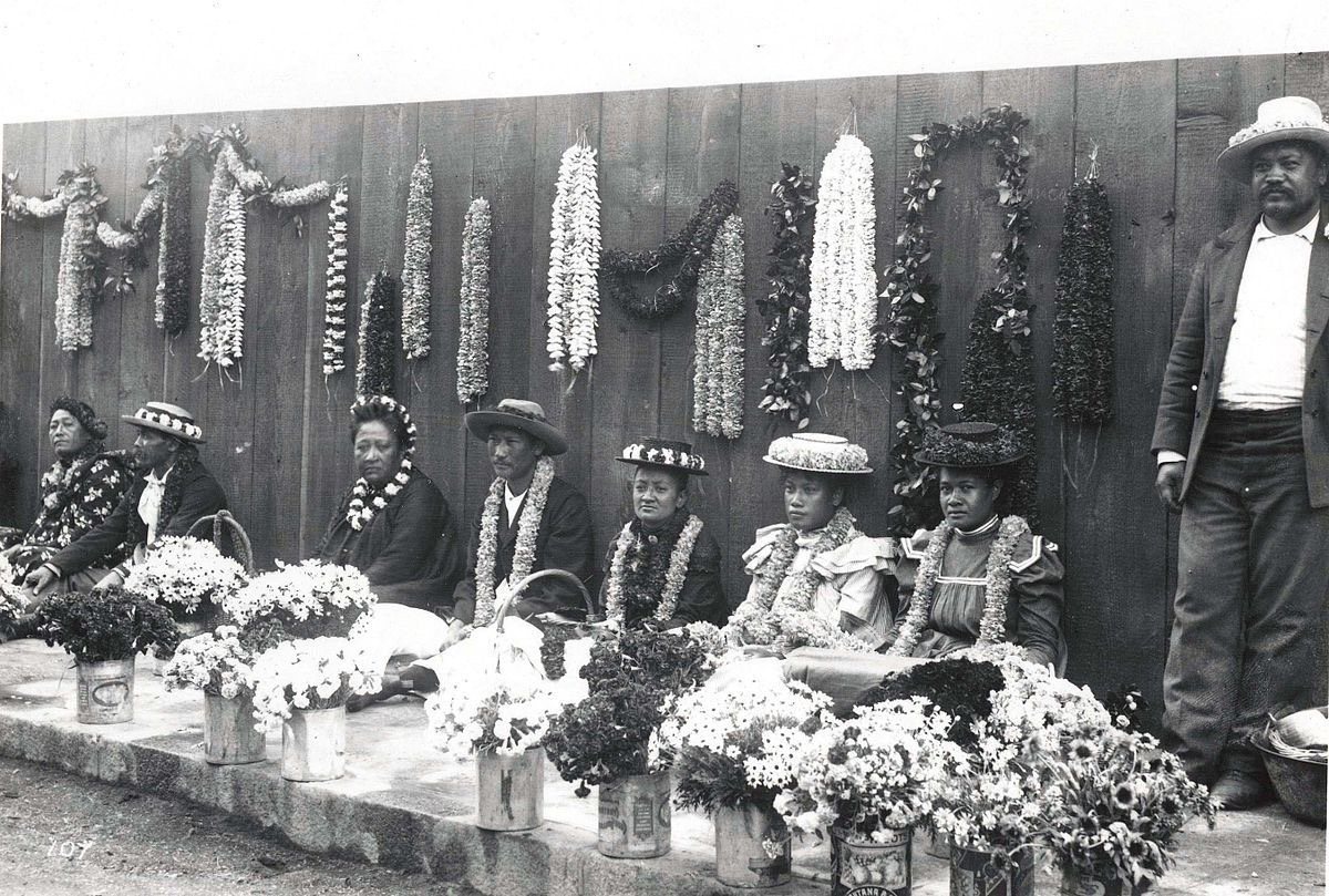 Black-and-white photograph of Hawaiian women seated on a Honolulu sidewalk with long strands of lei for sale, around 1900