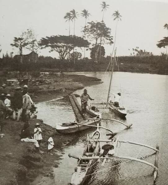 Black-and-white photograph of outrigger canoes moored at a river landing, with people gathered on the muddy bank and tall palms behind, late 1800s