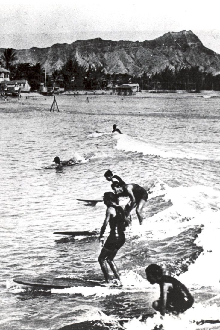 Black-and-white photograph of several surfers riding a small wave on long wooden boards at Waikīkī, with Diamond Head behind, early 1900s