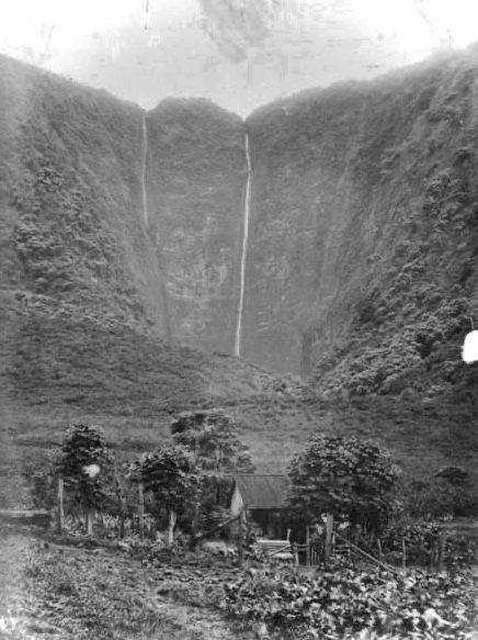 Black and white photograph of twin waterfalls dropping down a sheer valley wall above a small house and gardens, late 1800s